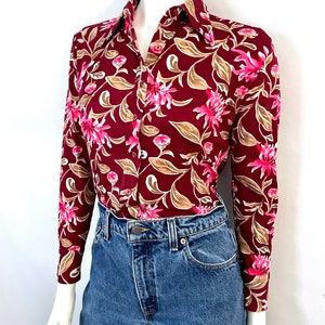 May include: A long-sleeved button-down shirt with a burgundy background and a floral pattern in pink, tan, and white. The shirt is paired with blue denim jeans.