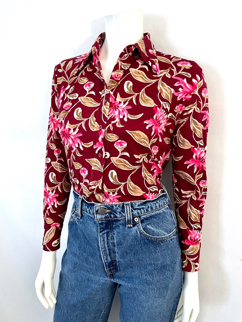May include: A long-sleeved button-down shirt with a burgundy background and a floral pattern in pink, tan, and white. The shirt is paired with blue denim jeans.
