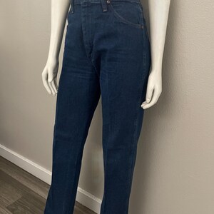 May include: Dark blue denim jeans with a high waist and a classic straight leg fit. The jeans feature a button and zipper closure, front and back pockets, and a copper-colored button. The jeans are displayed on a mannequin.