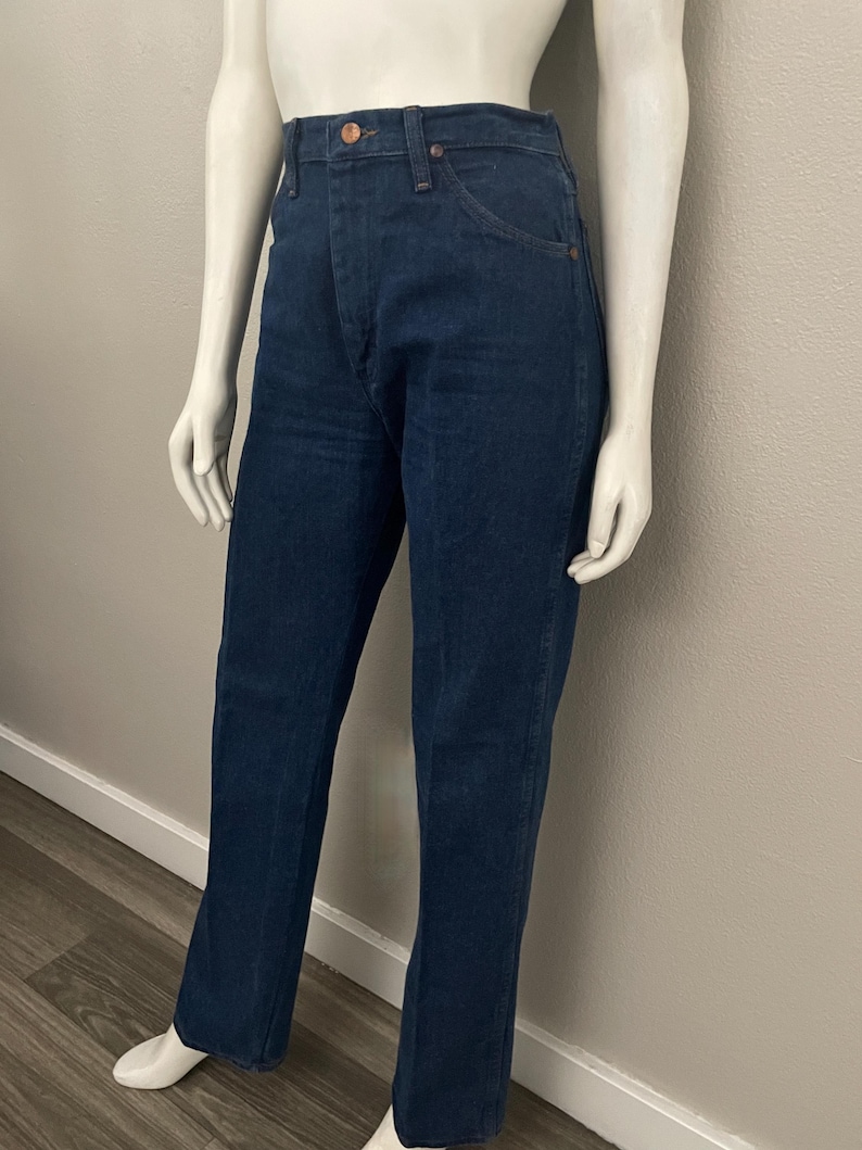 May include: Dark blue denim jeans with a high waist and a classic straight leg fit. The jeans feature a button and zipper closure, front and back pockets, and a copper-colored button. The jeans are displayed on a mannequin.