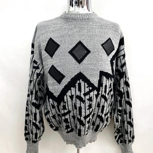 May include: A gray sweater with a geometric design. The sweater features black diamond shapes and a black and gray pattern on the sleeves and lower portion. The sweater is displayed on a mannequin.