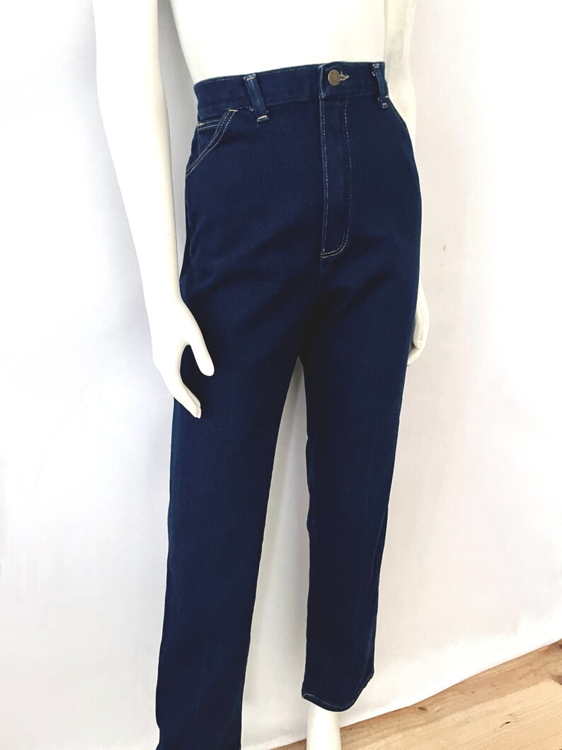 K&ouml;nnte beinhalten: Dunkelblaue Jeans mit hoher Taille und geradem Bein. Die Jeans hat einen Knopf- und Rei&szlig;verschluss und sichtbare N&auml;hte in einer hellen Farbe. Die Jeans wird auf einer Schaufensterpuppe pr&auml;sentiert.