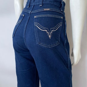 K&ouml;nnte beinhalten: Dunkelblaue Wrangler-Jeans mit hohem Bund. Die Ges&auml;&szlig;taschen haben ein markantes wei&szlig;es Stickdesign. Das Wrangler-Logo ist &uuml;ber der rechten Ges&auml;&szlig;tasche sichtbar. Die Jeans haben eine klassische, gerade Beinpassform.