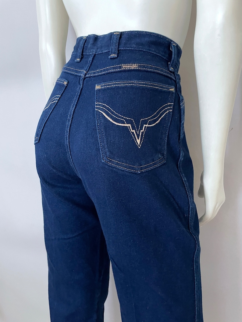 K&ouml;nnte beinhalten: Dunkelblaue Wrangler-Jeans mit hohem Bund. Die Ges&auml;&szlig;taschen haben ein markantes wei&szlig;es Stickdesign. Das Wrangler-Logo ist &uuml;ber der rechten Ges&auml;&szlig;tasche sichtbar. Die Jeans haben eine klassische, gerade Beinpassform.