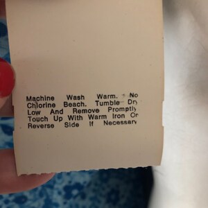 May include: A close-up of a white tag with care instructions. The tag has a hole at the top and text that reads: Machine Wash, Chlorine Beach, Low, Touch Up With Warm Iron Or Reverse Side If Necessary.
