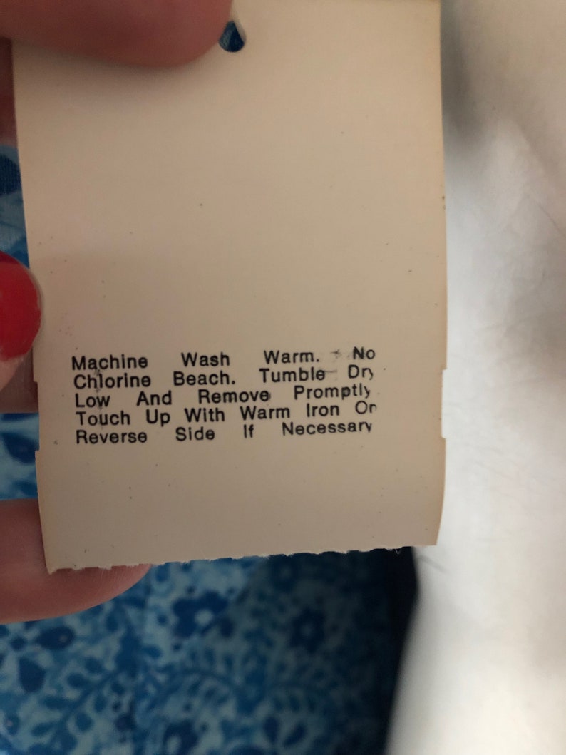 May include: A close-up of a white tag with care instructions. The tag has a hole at the top and text that reads: Machine Wash, Chlorine Beach, Low, Touch Up With Warm Iron Or Reverse Side If Necessary.