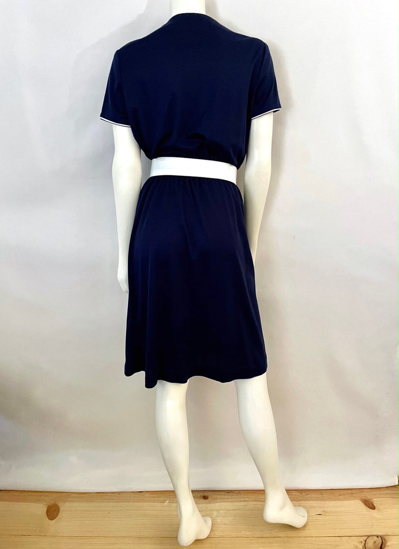 Op de afbeelding: Marineblauwe jurk met een witte riem. De jurk heeft korte mouwen met witte bies en een V-hals. De rok is A-lijn. De jurk wordt op een mannequin gepresenteerd.