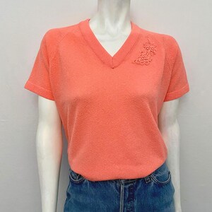 May include: Coral-colored V-neck short-sleeve shirt with a textured surface. A floral design is on the upper left chest. The shirt is paired with blue denim jeans.