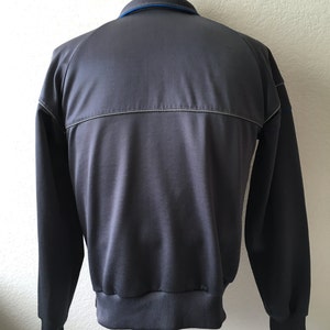 May include: A grey track jacket with a blue collar and a silver stripe across the back. The jacket has long sleeves and a ribbed hem and cuffs. The jacket is displayed on a mannequin.