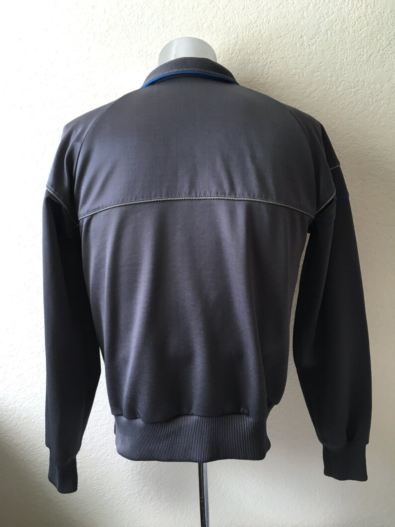 May include: A grey track jacket with a blue collar and a silver stripe across the back. The jacket has long sleeves and a ribbed hem and cuffs. The jacket is displayed on a mannequin.