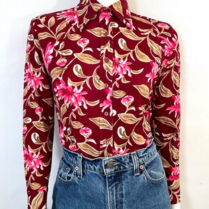 May include: Burgundy long-sleeve button-up shirt with a floral pattern. The shirt features pink flowers, tan leaves, and white accents. The shirt is paired with blue denim jeans.