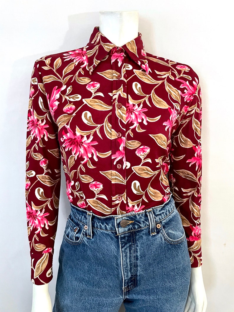 May include: Burgundy long-sleeve button-up shirt with a floral pattern. The shirt features pink flowers, tan leaves, and white accents. The shirt is paired with blue denim jeans.
