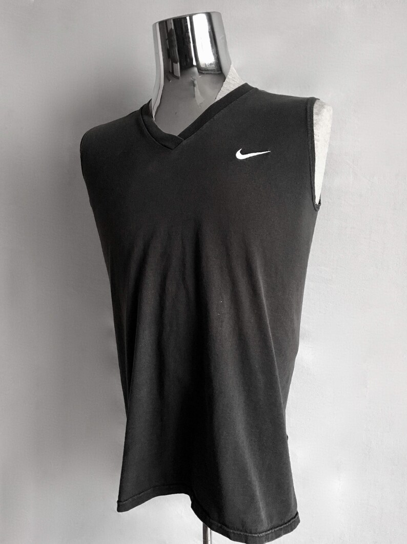 Puede incluir: Una camiseta sin mangas gris oscuro con cuello en V. La camiseta presenta un peque&ntilde;o logotipo blanco de Nike en la parte superior izquierda del pecho. La prenda se exhibe en un maniqu&iacute;.