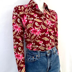 May include: A long-sleeved button-down shirt with a floral pattern. The shirt features a deep burgundy background with pink and white flowers and tan leaves. The shirt is paired with blue denim jeans.