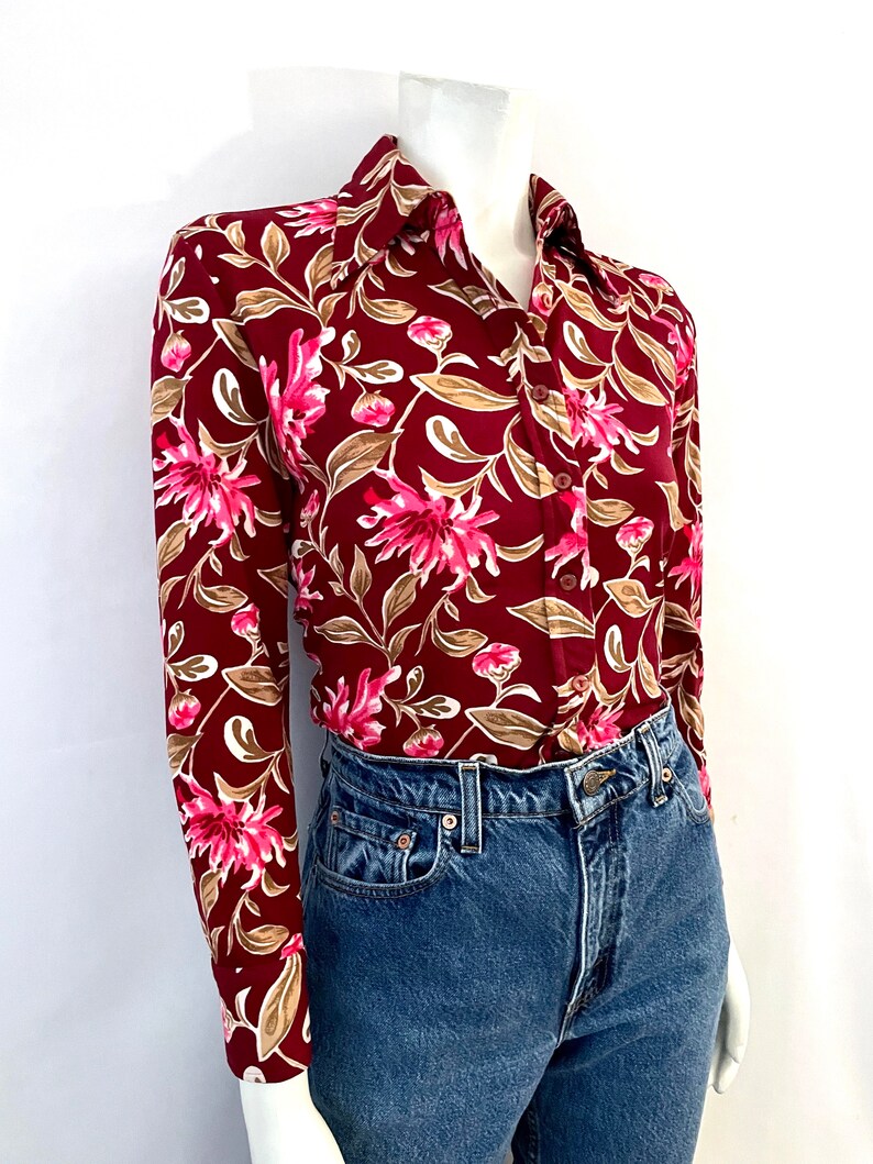 May include: A long-sleeved button-down shirt with a floral pattern. The shirt features a deep burgundy background with pink and white flowers and tan leaves. The shirt is paired with blue denim jeans.