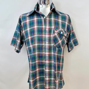 Puede incluir: Una camisa a cuadros de manga corta en un patrón clásico de verde, rojo, blanco y azul. La camisa tiene una botonadura frontal, un bolsillo en el pecho y un cuello. La camisa se exhibe en un maniquí.
