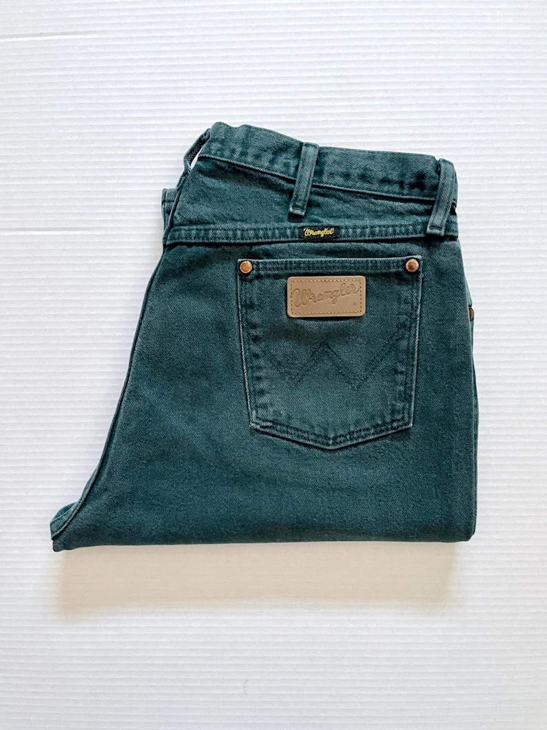 May include: Folded dark teal Wrangler jeans. The jeans feature a classic five-pocket design, belt loops, and a brown leather patch with the Wrangler logo on the back pocket. The jeans are made of a durable denim material.