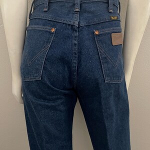 May include: Dark blue denim jeans with a classic five-pocket design. Features copper rivets, a brown leather patch, and the Wrangler logo. The jeans have a straight leg cut and a high waist. The back pockets have a distinctive stitching pattern.