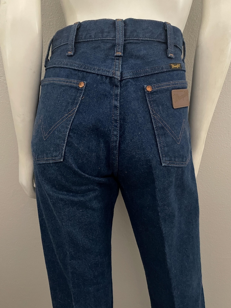 May include: Dark blue denim jeans with a classic five-pocket design. Features copper rivets, a brown leather patch, and the Wrangler logo. The jeans have a straight leg cut and a high waist. The back pockets have a distinctive stitching pattern.