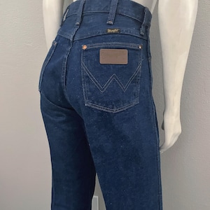 May include: Dark blue denim jeans with a high waist. The jeans feature the Wrangler logo on the back pocket and a brown leather patch. The stitching is a contrasting light color. The jeans are displayed on a mannequin.