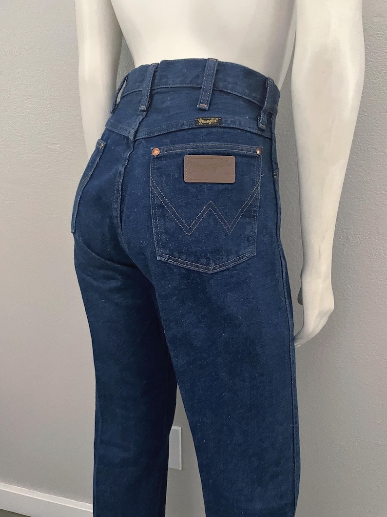 May include: Dark blue denim jeans with a high waist. The jeans feature the Wrangler logo on the back pocket and a brown leather patch. The stitching is a contrasting light color. The jeans are displayed on a mannequin.