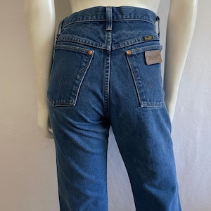 May include: Dark blue denim jeans with a high waist and a straight leg. The back pockets have a classic design, and a brown leather patch with the Wrangler logo is visible. The jeans are displayed on a mannequin.