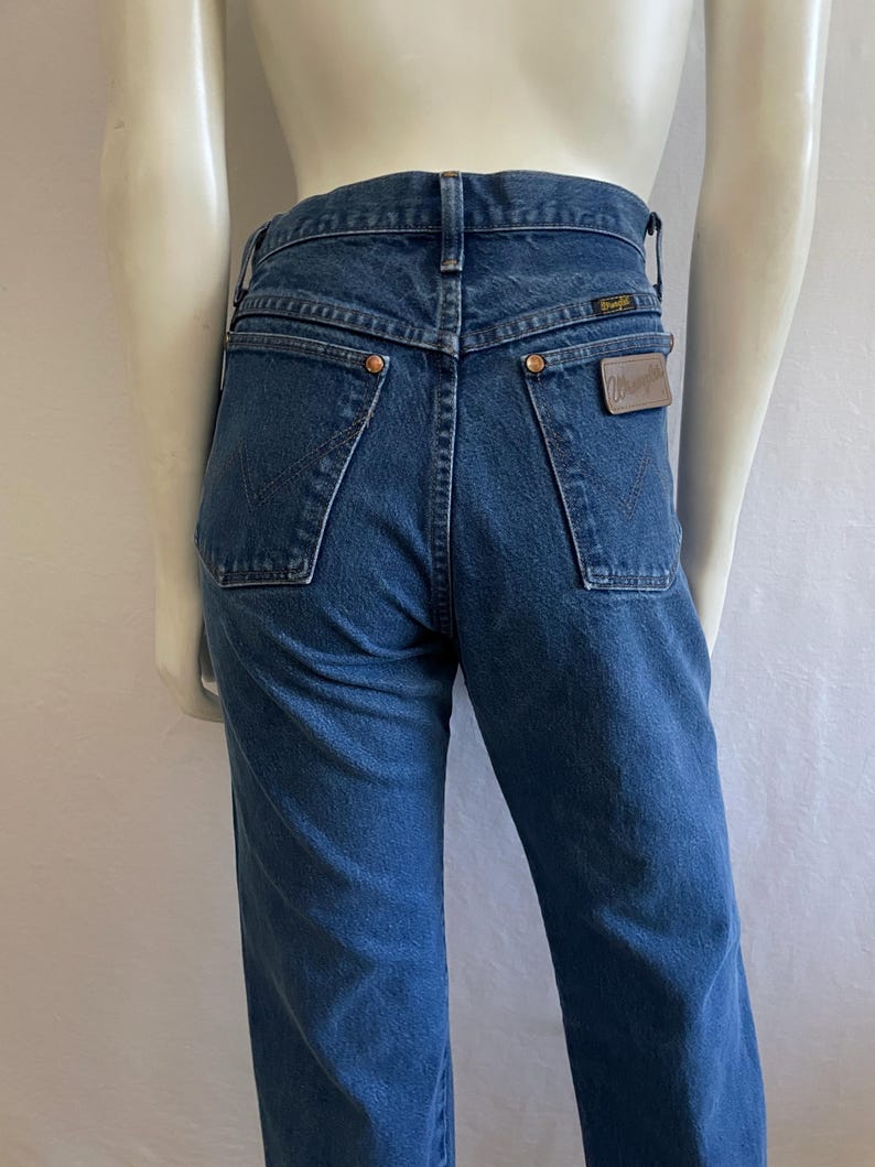 May include: Dark blue denim jeans with a high waist and a straight leg. The back pockets have a classic design, and a brown leather patch with the Wrangler logo is visible. The jeans are displayed on a mannequin.