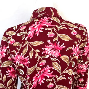 May include: A burgundy button-down shirt with a vibrant floral pattern. The design features large pink flowers, tan leaves, and smaller pink buds against a dark red background. The shirt's collar is visible.