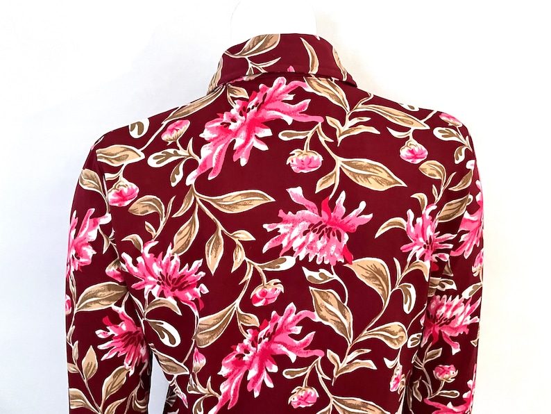 May include: A burgundy button-down shirt with a vibrant floral pattern. The design features large pink flowers, tan leaves, and smaller pink buds against a dark red background. The shirt's collar is visible.