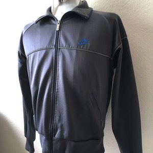 May include: A dark grey track jacket with a full zip, a blue Nike logo, and blue trim on the collar. The jacket features a ribbed hem and cuffs, and two small pockets. The jacket is a classic sportswear item.