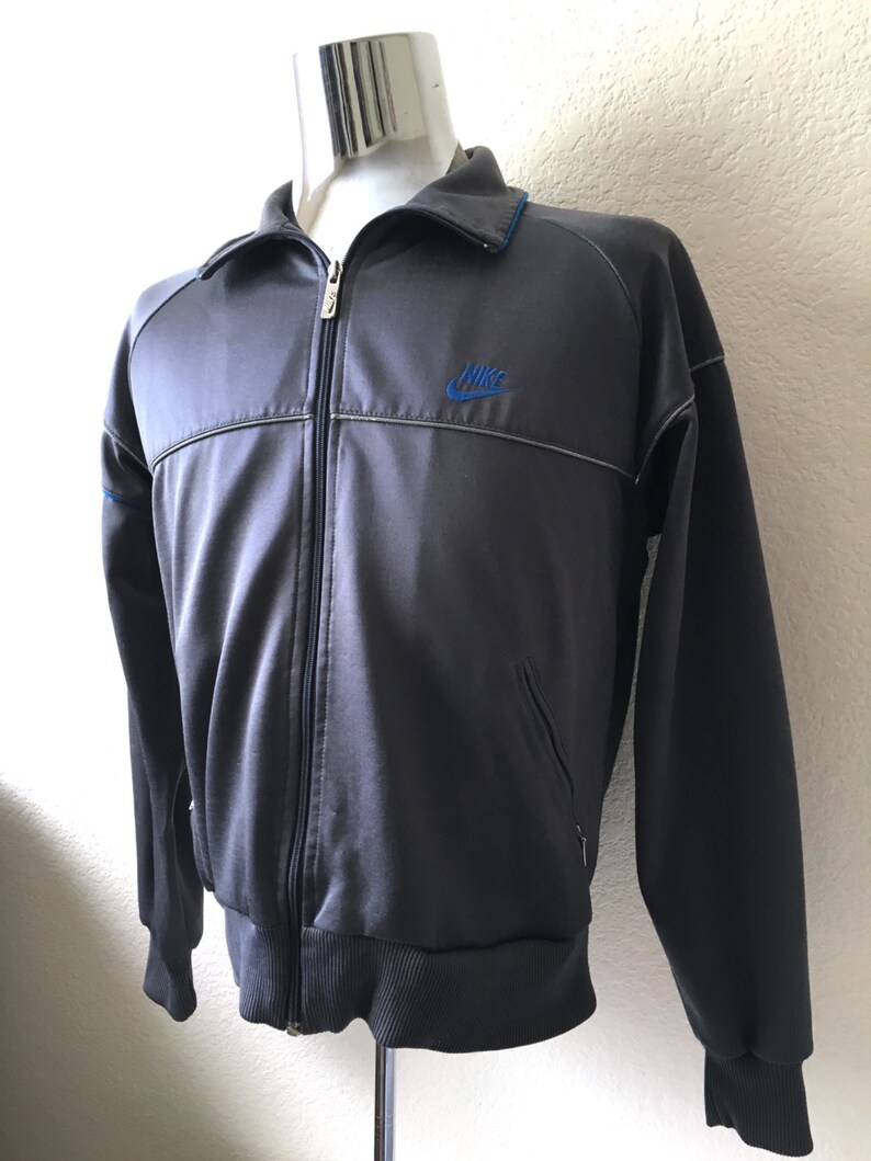 May include: A dark grey track jacket with a full zip, a blue Nike logo, and blue trim on the collar. The jacket features a ribbed hem and cuffs, and two small pockets. The jacket is a classic sportswear item.