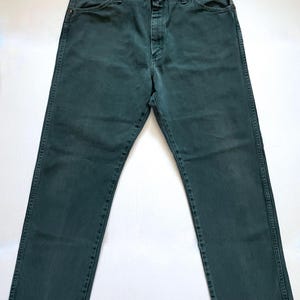 May include: Dark teal denim jeans with a classic five-pocket design. The jeans have a button and zipper closure, and copper-colored rivets. The denim appears to be pre-washed, giving it a slightly faded look. The jeans are laid flat on a white surface.