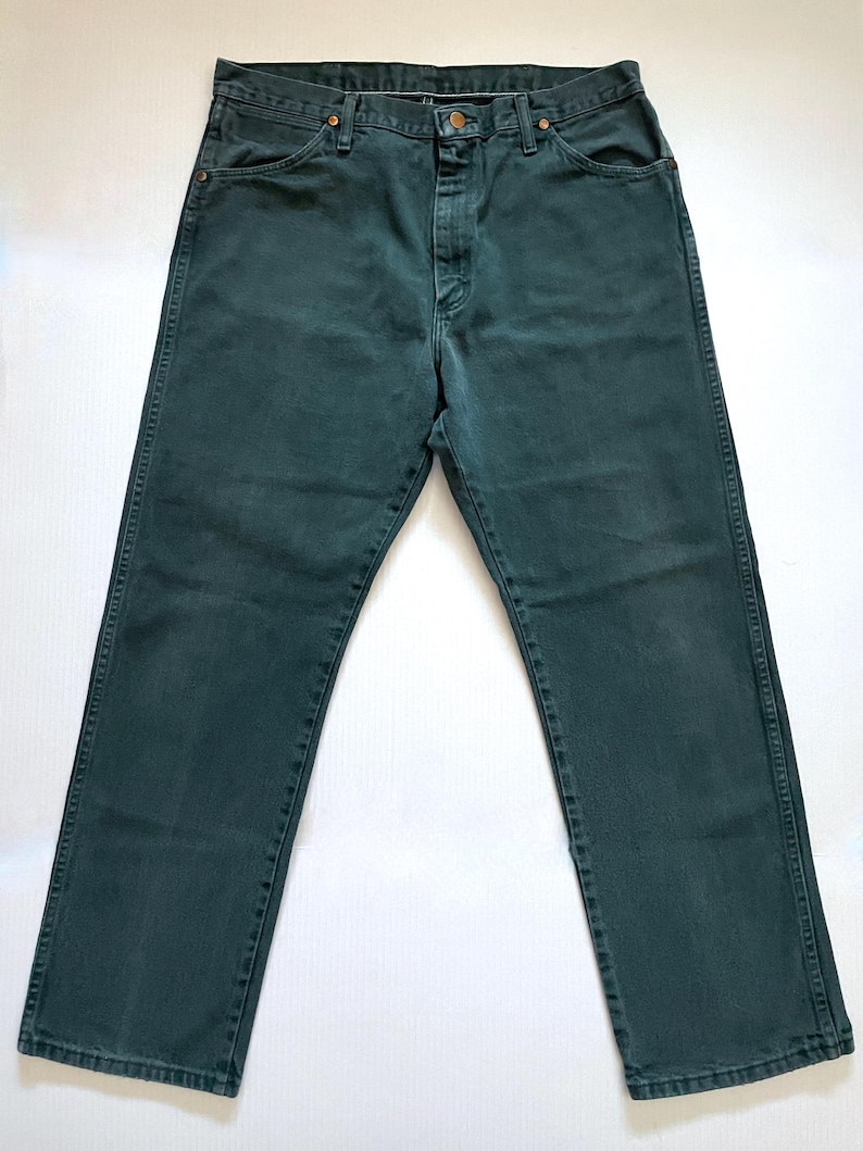 May include: Dark teal denim jeans with a classic five-pocket design. The jeans have a button and zipper closure, and copper-colored rivets. The denim appears to be pre-washed, giving it a slightly faded look. The jeans are laid flat on a white surface.