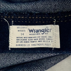 K&ouml;nnte beinhalten: Nahaufnahme von dunkelblauem Denim-Stoff mit einem wei&szlig;en Etikett. Das Etikett tr&auml;gt die Aufschrift "Wrangler" und enth&auml;lt die Gr&ouml;&szlig;e 14, Pflegehinweise und "Made in U.S.A."