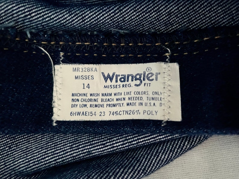 K&ouml;nnte beinhalten: Nahaufnahme von dunkelblauem Denim-Stoff mit einem wei&szlig;en Etikett. Das Etikett tr&auml;gt die Aufschrift "Wrangler" und enth&auml;lt die Gr&ouml;&szlig;e 14, Pflegehinweise und "Made in U.S.A."