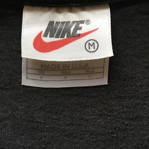 Puede incluir: Primer plano de una camiseta negra con una etiqueta blanca. La etiqueta presenta el logotipo de Nike en rojo y blanco, con la palabra "NIKE" y una "R" en c&iacute;rculo. Debajo, el texto "MADE IN U.S.A." e informaci&oacute;n de tallas.