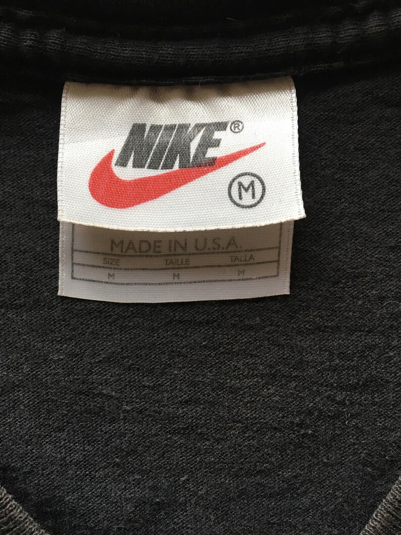 Puede incluir: Primer plano de una camiseta negra con una etiqueta blanca. La etiqueta presenta el logotipo de Nike en rojo y blanco, con la palabra "NIKE" y una "R" en c&iacute;rculo. Debajo, el texto "MADE IN U.S.A." e informaci&oacute;n de tallas.