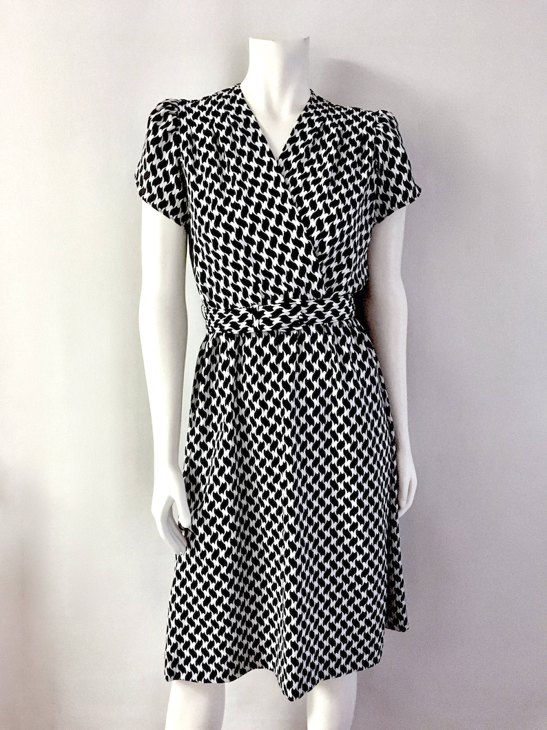 May include: A black and white patterned dress with a wrap-style design. The dress features short sleeves and a V-neckline. A matching belt cinches the waist. The fabric has a repeating pattern of black shapes on a white background.