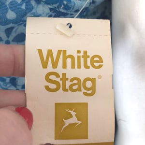 May include: A close-up of a White Stag clothing tag. The tag is cream-colored with the words "White Stag" in gold lettering. Below the text is a gold square with a white deer silhouette. The tag is attached to a blue floral patterned fabric.