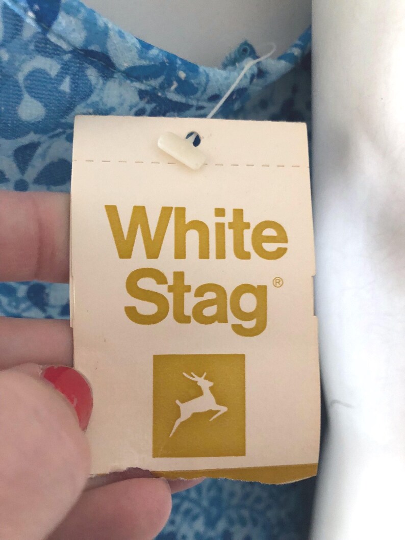 May include: A close-up of a White Stag clothing tag. The tag is cream-colored with the words "White Stag" in gold lettering. Below the text is a gold square with a white deer silhouette. The tag is attached to a blue floral patterned fabric.