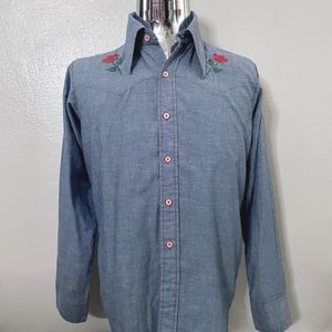 May include: A blue long-sleeve shirt with red rose embroidery on the collar. The shirt has a button-down front with pink buttons and stitching. The shirt is displayed on a mannequin.
