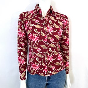 May include: A long-sleeved button-down shirt with a burgundy background and a floral pattern in pink and tan. The shirt is displayed on a mannequin and paired with blue jeans.