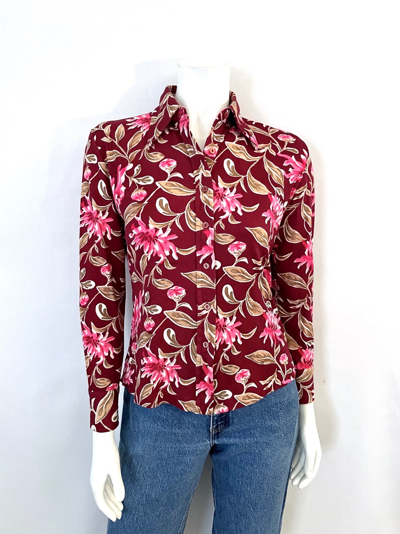May include: A long-sleeved button-down shirt with a burgundy background and a floral pattern in pink and tan. The shirt is displayed on a mannequin and paired with blue jeans.