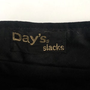 May include: Close-up of a black fabric with a tag that reads "Day's slacks" in gold lettering. The tag is rectangular and sewn onto the fabric. The fabric appears to be a dark, possibly corduroy, material.