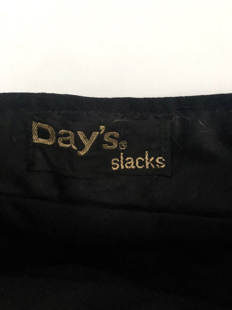 May include: Close-up of a black fabric with a tag that reads "Day's slacks" in gold lettering. The tag is rectangular and sewn onto the fabric. The fabric appears to be a dark, possibly corduroy, material.