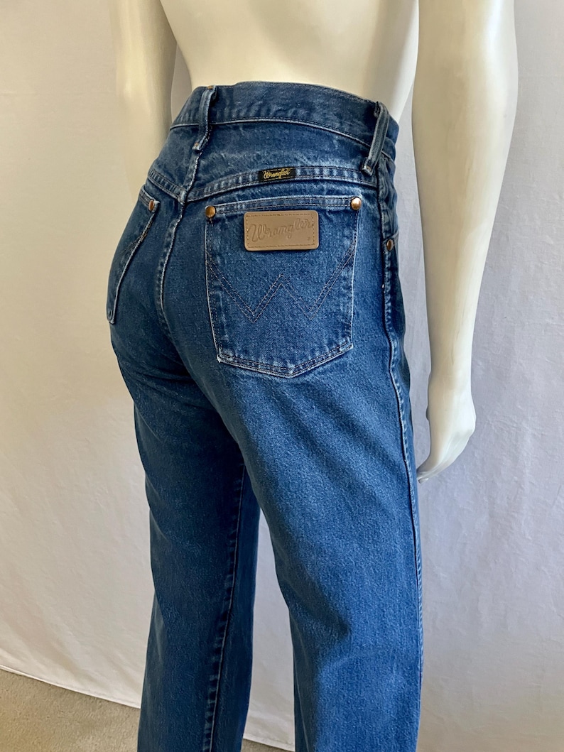 May include: Blue denim jeans with a high waist and a classic five-pocket design. The back features a leather patch with the "Wrangler" logo and a stitched "W" on the back pockets. The jeans are displayed on a mannequin.