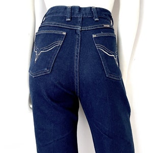 K&ouml;nnte beinhalten: Dunkelblaue Jeans aus Denim mit wei&szlig;en Ziern&auml;hten. Die Jeans hat eine hohe Taille und ein klassisches F&uuml;nf-Taschen-Design. Die Ges&auml;&szlig;taschen haben ein dekoratives Nahtmuster. Die Jeans wird auf einer Schaufenquin pr&auml;sentiert.