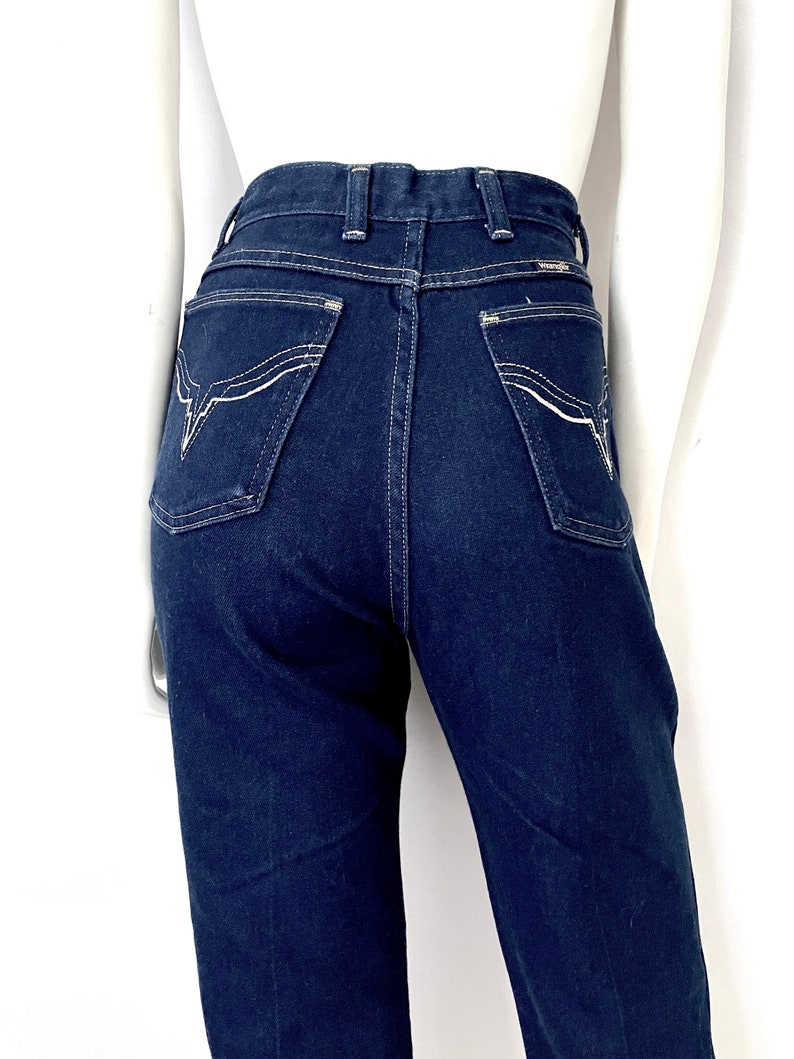 K&ouml;nnte beinhalten: Dunkelblaue Jeans aus Denim mit wei&szlig;en Ziern&auml;hten. Die Jeans hat eine hohe Taille und ein klassisches F&uuml;nf-Taschen-Design. Die Ges&auml;&szlig;taschen haben ein dekoratives Nahtmuster. Die Jeans wird auf einer Schaufenquin pr&auml;sentiert.