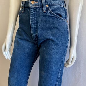 May include: Blue denim jeans with a classic five-pocket design. The jeans feature a button and zipper closure, belt loops, and copper-colored rivets. The denim has a medium wash with subtle fading and wear.