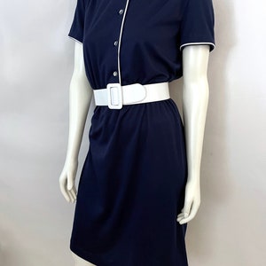 Op de afbeelding: Een marineblauwe jurk met witte bies en bijpassende witte riem. De jurk met korte mouwen heeft een knoopsluiting aan de voorkant en een zoom tot op de knie. De jurk wordt op een mannequin getoond.